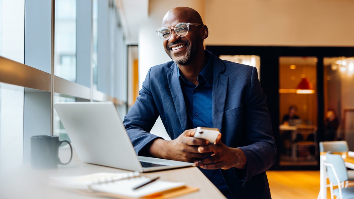 A confident adult male wearing a formal suit, smiling while engaged with a laptop and smartphone. The setting is bright and comfortable, suggesting productivity, focus, and a positive demeanor.